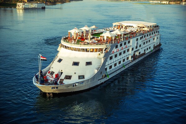 Un gran barco de crucero fluvial con varias cubiertas, zonas de estar al aire libre y una bandera en la proa.