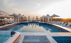 Un área de piscina con sillones de playa y sombrillas, un jacuzzi y vistas a la puesta de sol sobre el agua.