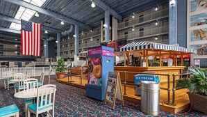 Een ruime binnenruimte met een vloerkleed met patroon, een grote Amerikaanse vlag, een kiosk voor donuts en een ijsstand.