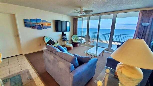 Een woonkamer met een bank, een tafel met een lamp, een televisie, en een balkon met uitzicht op de oceaan.