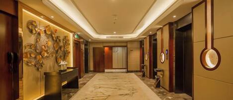 A hotel lobby with a marble floor, a reception desk, a wall with decorative art, and an elevator.