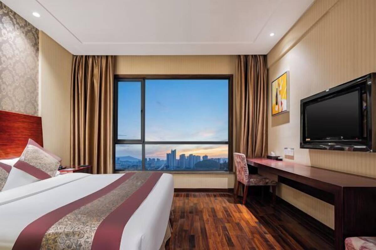 Une chambre d’hôtel avec un grand lit, un bureau, une chaise, une télévision et une vue sur le paysage urbain, visible à travers une grande fenêtre.