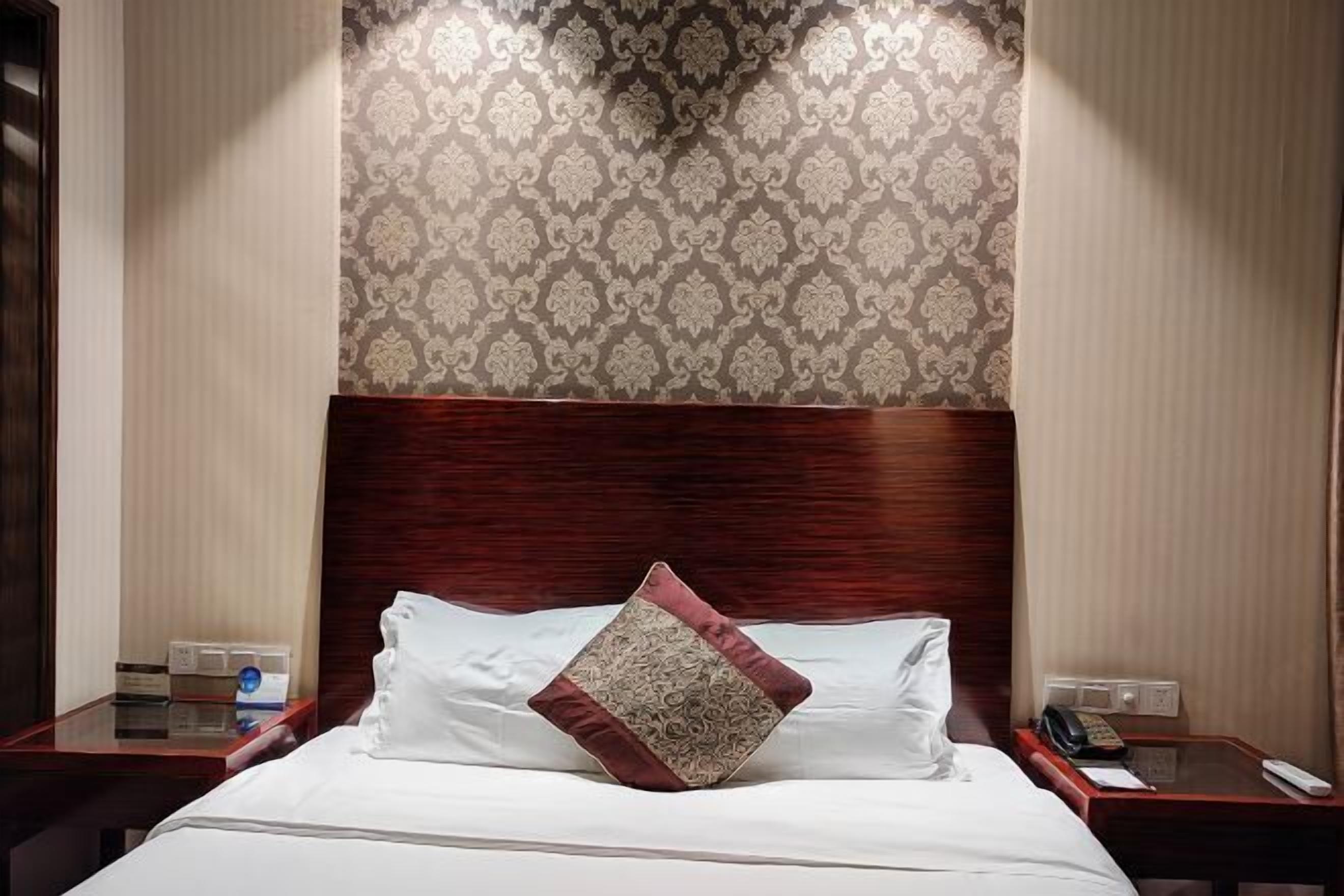 A hotel room with a large bed, a wooden headboard, a patterned wallpaper, and a nightstand with a telephone and a remote control.