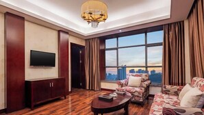 A modern living room with a flat-screen TV, a wooden coffee table, floral-patterned sofas, a large window with a city view, and a chandelier. - Howard Johnson Park Plaza Hotel (Jingshan Railway Station Inn) (Jingshan)