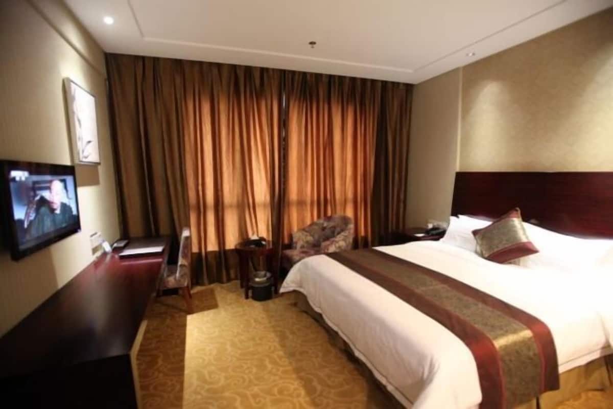 A hotel room with a large bed, a flat-screen TV mounted on the wall, a desk with a chair, and a large window with curtains.