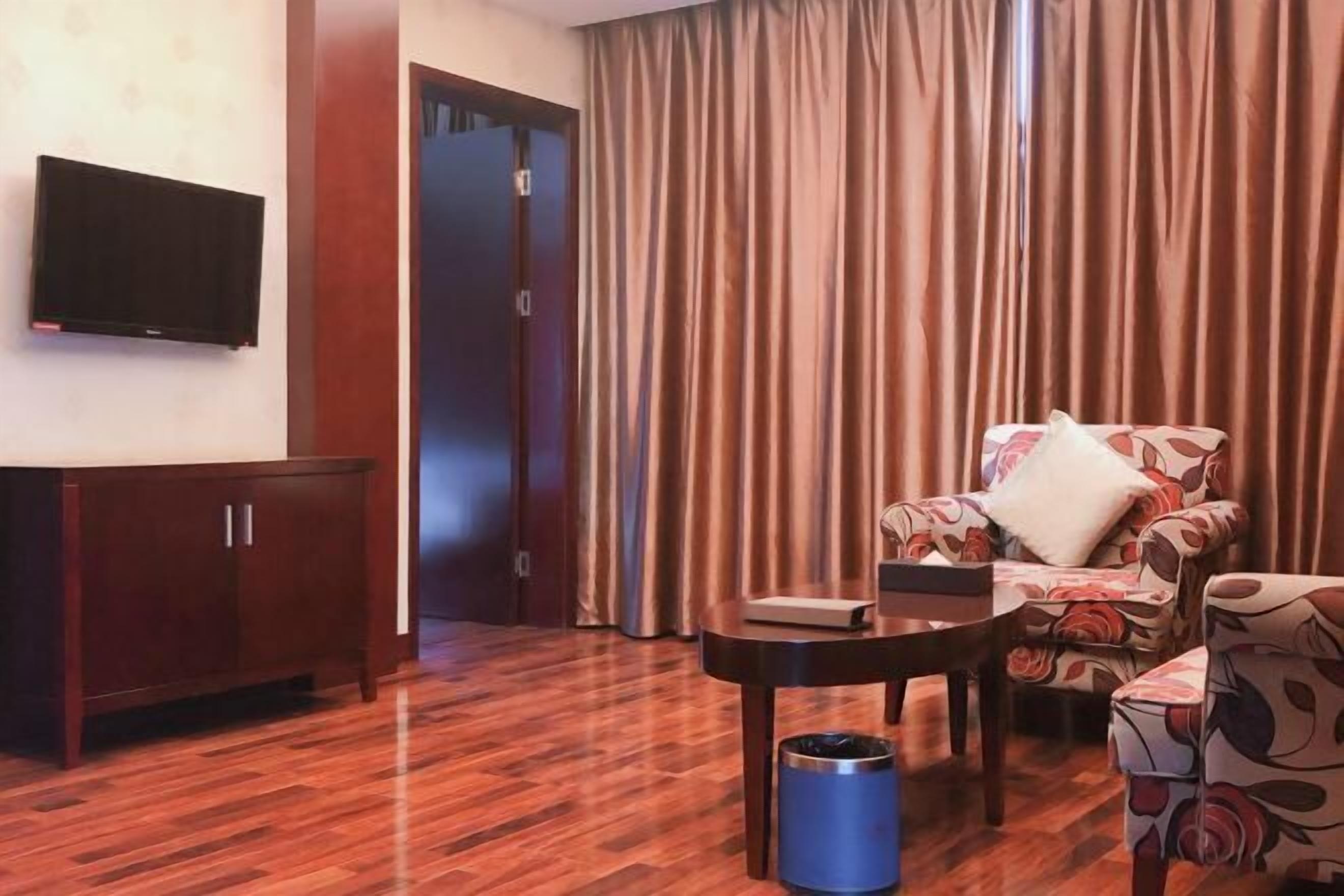 Habitación de hotel con piso de madera, televisor montado en la pared, sofá con tapicería floral, una mesita y un bote de basura azul.