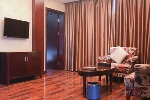 A hotel room with a wooden floor, a TV mounted on the wall, a sofa with floral upholstery, a small table, and a blue trash bin. - Howard Johnson Park Plaza Hotel (Jingshan Railway Station Inn) (Jingshan)