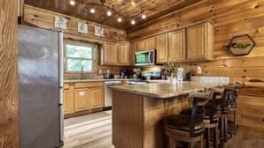 A kitchen with wooden cabinets, a refrigerator, a microwave, a granite countertop, and bar stools. - Cozy Bear Lodge-peaceful, easy access, beautiful view (Sevierville)