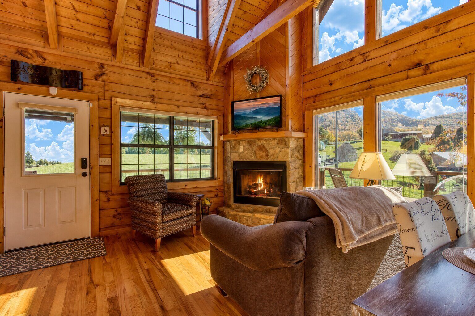 L’intérieur d’une cabane en rondins, avec une cheminée en pierre, un canapé, un fauteuil à motifs et de grandes fenêtres offrant une vue panoramique.
