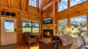 A log cabin interior with a stone fireplace, a couch, a patterned armchair, and large windows offering scenic views. - Picture Perfect , Panoramic Mountain Views, $560 FREE tickets each paid day! (Sevierville)