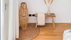 A room with a wooden floor, a woven rug, a wicker chair, a small wooden table with a vase of pampas grass, and a straw hat on the wall.