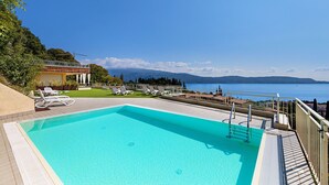 A rectangular swimming pool with a clear blue water, surrounded by a deck with lounge chairs and a view of the sea and mountains in the background.