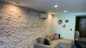 A living room with a stone wall, a beige sectional sofa, a blue chair, and a small table. - Super Chic 3bd aptm. sauna/jacuzzi Mamitas Beach (Playa del Carmen)