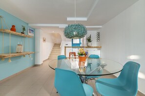 A modern kitchen with a glass dining table, teal chairs, and a hanging light fixture.
