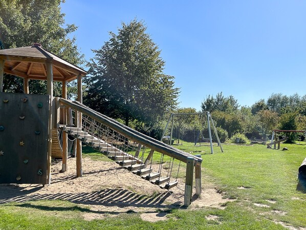 Een speeltuin met een houten klimconstructie, schommel en een overdekte zitplek.