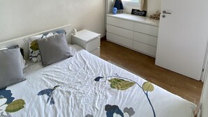 Une chambre à coucher comprenant un lit, des tables de chevet, une commode, une fenêtre avec des rideaux, un radiateur et un réfrigérateur.