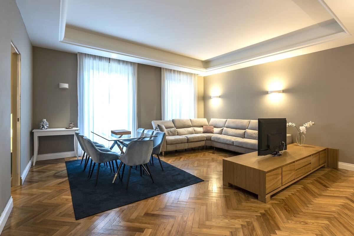 Una sala moderna con suelo de madera en espiga, un sofá modular, una zona de comedor con mesa y sillas de vidrio, un televisor sobre un mueble de madera y un amplio ventanal con cortinas.