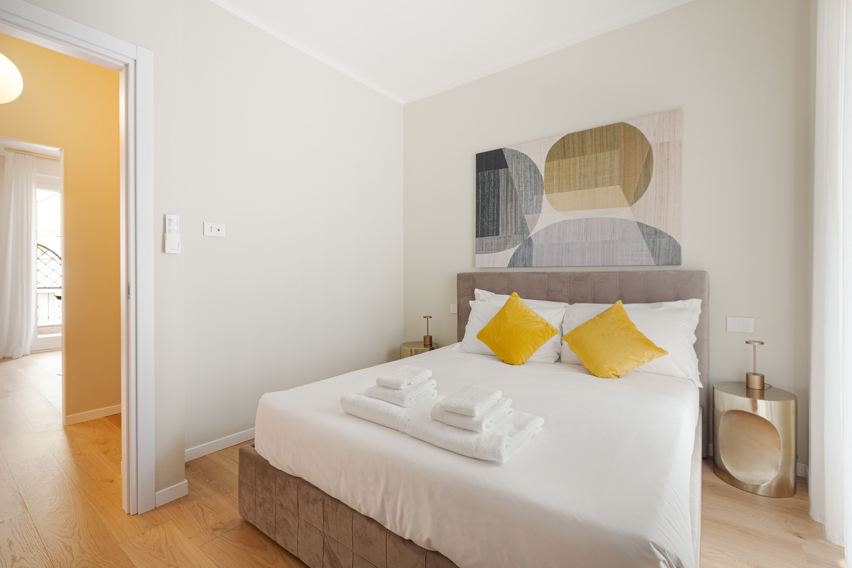 A neatly made bed with white linens and two yellow pillows, a bedside table with a lamp, and a modern abstract painting on the wall.