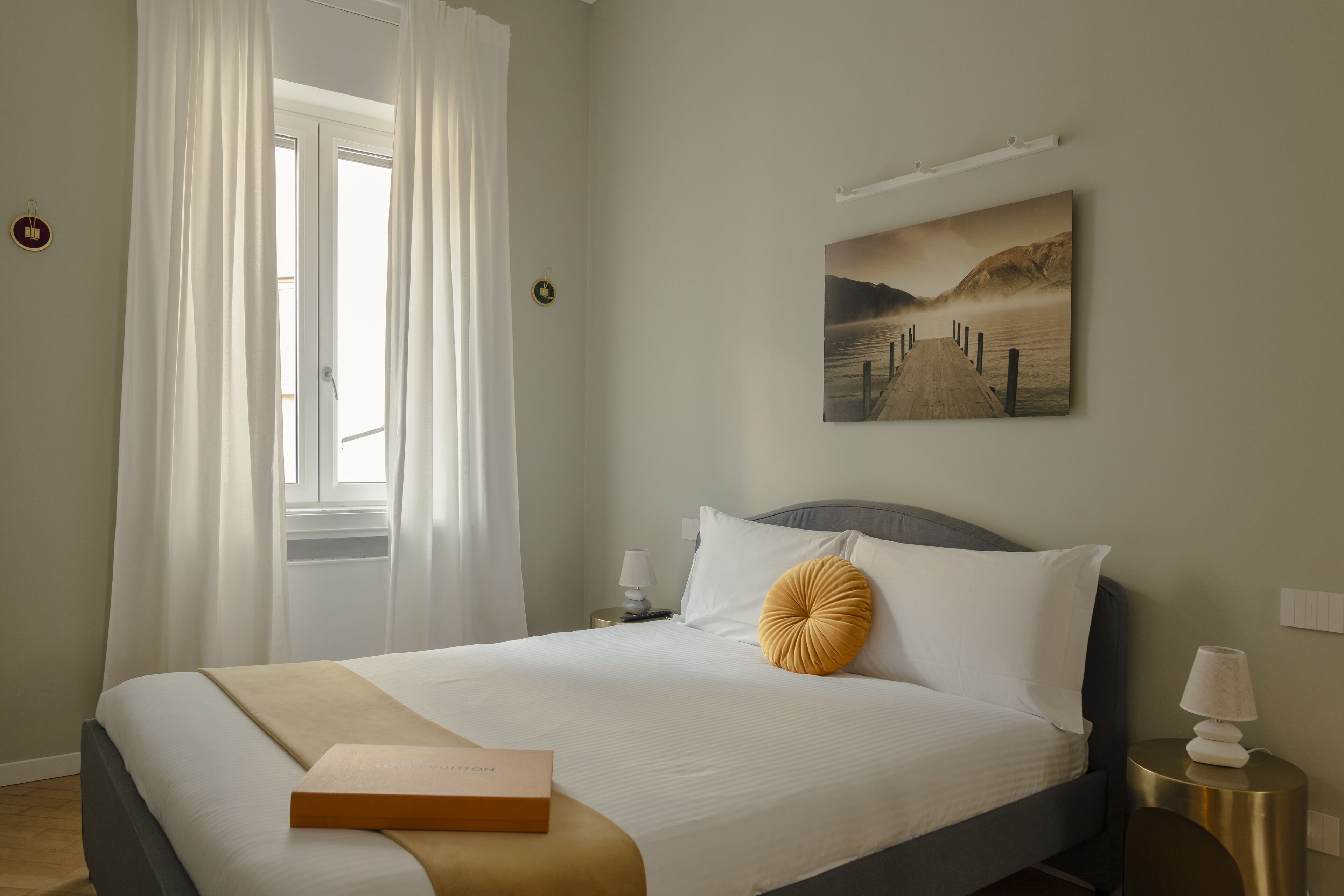 Una cama bien hecha con edredón blanco y una sola almohada decorativa, una mesita de noche con lámpara, un cuadro en la pared y una ventana con cortinas blancas.