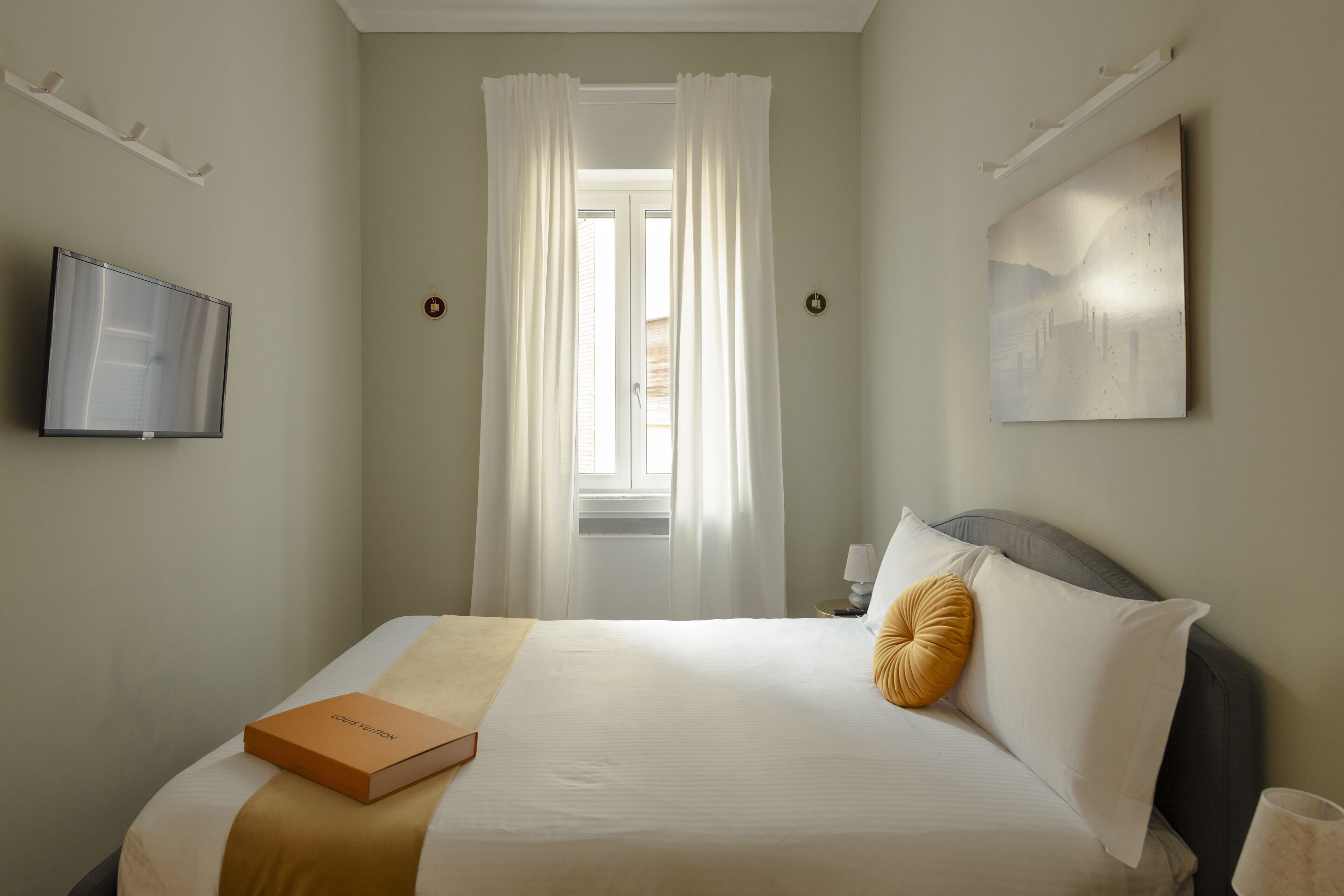 A neatly made bed with a book, a television on the wall, and a painting.