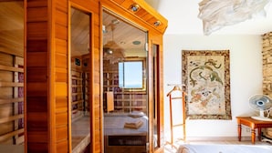 Une pièce avec une armoire en bois, un lit recouvert de draps blancs soigneusement pliés, un ventilateur fixé au mur et une tapisserie traditionnelle.