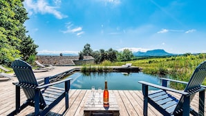 Une terrasse en bois avec deux fauteuils Adirondack, une bouteille de vin et deux verres sur une table, donnant sur une étendue d’eau calme et un paysage montagneux.