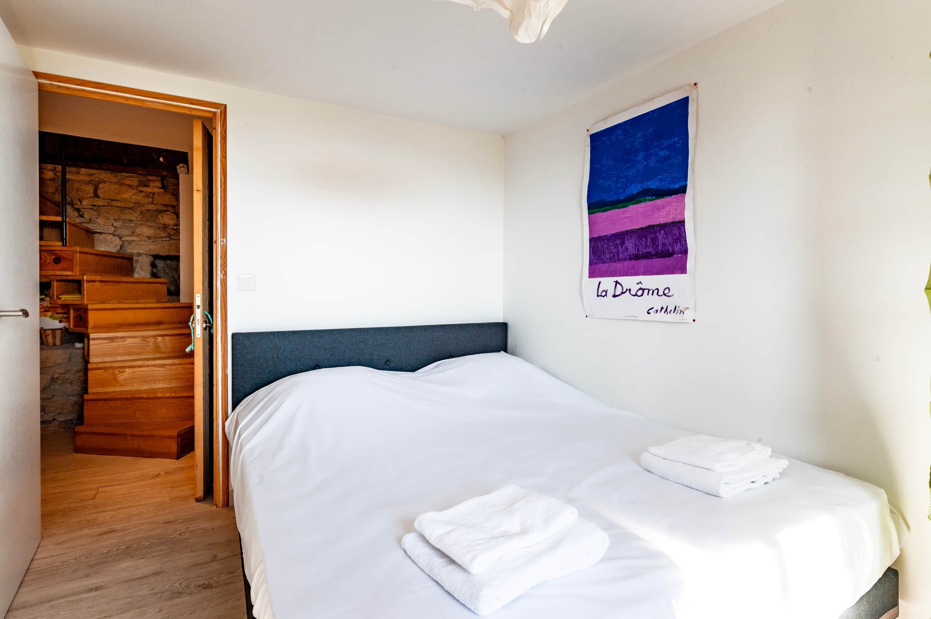 Une chambre à coucher avec un lit, une porte en bois donnant sur un escalier et un mur orné d’un tableau.