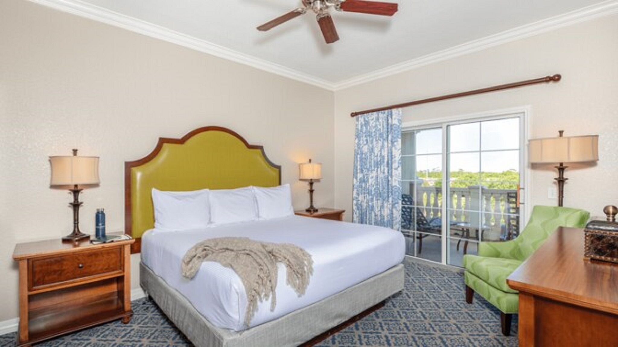 A hotel room with a bed, two bedside tables, a ceiling fan, a green armchair, and a view through a glass door.