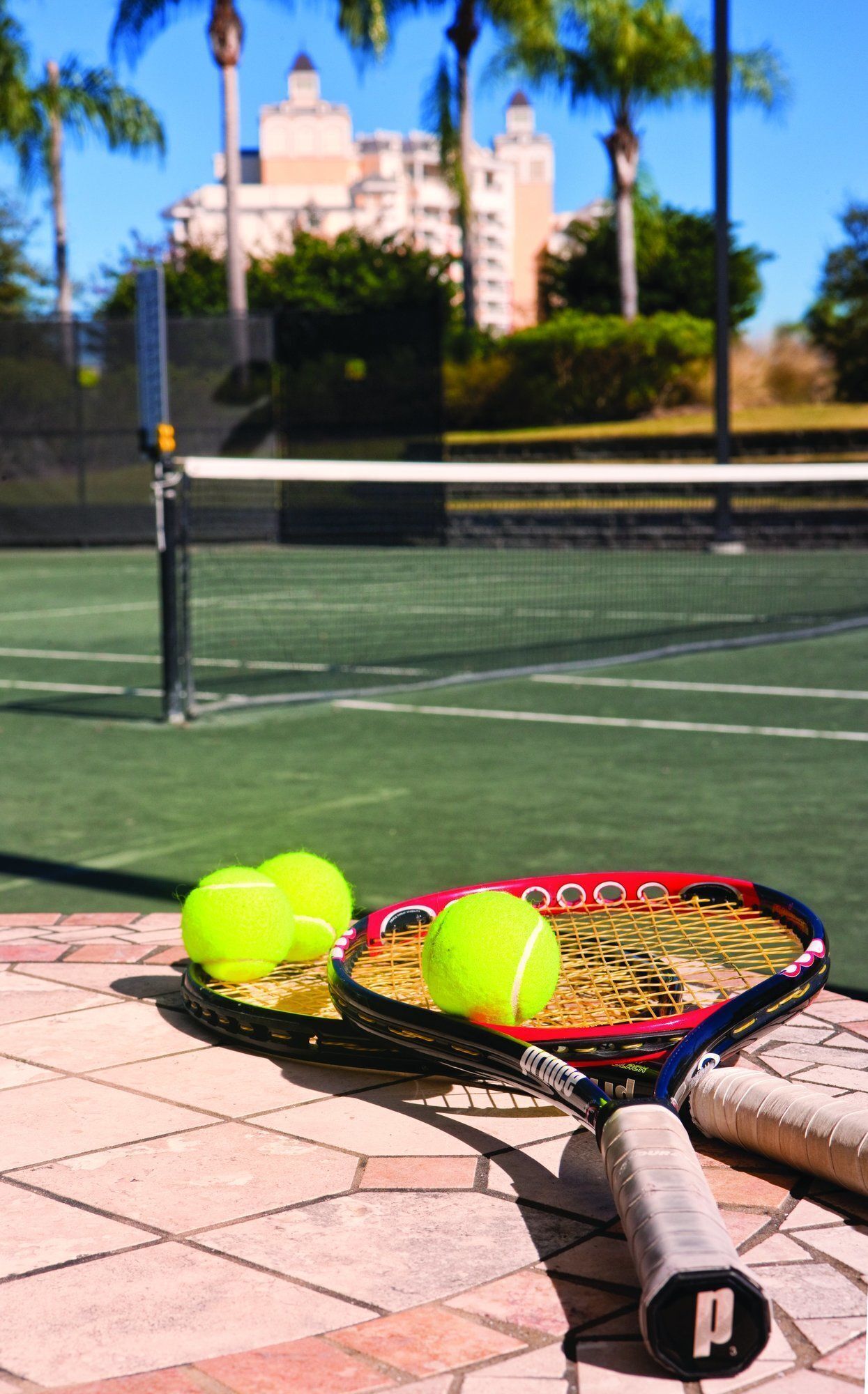 Una cancha de tenis con red, tres pelotas y una raqueta sobre una superficie de baldosas.
