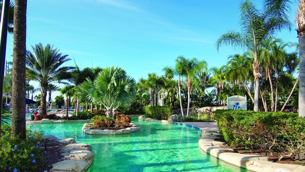 A clear pool surrounded by palm trees and tropical plants, with a pathway leading to a building. - Worldmark Reunion (Kissimmee)