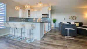 A modern kitchen with a central island, white cabinetry, and a bar stool area. There is a microwave, a potted plant, and a grey sofa in the living area. - Intervillas Florida - Villa Brightwater  (Cape Coral)