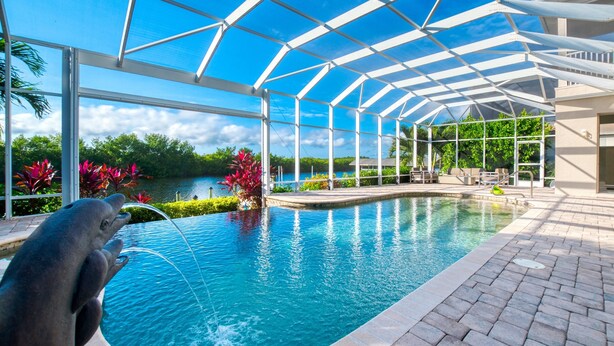 A swimming pool with a glass ceiling, surrounded by greenery and a view of a body of water. - Intervillas Florida - Villa Brightwater  (Cape Coral)