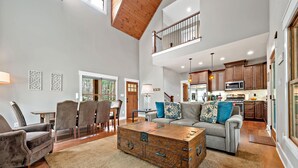 A spacious living room with a high ceiling, a dining area with chairs, a sofa with decorative pillows, and a kitchen with wooden cabinets and a microwave.