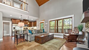 A spacious living area with a large wooden coffee table, a sofa, and a leather armchair. There is a kitchen in the background with modern appliances and a dining area.