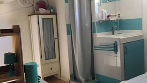 Une chambre à coucher de taille modeste, comprenant un lit, une petite table de chevet, une commode, et une salle de bain avec un lavabo, un miroir et un rideau de douche.