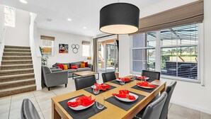 A dining area with a wooden table set for six, a staircase, a grey sofa, and a large window with blinds.