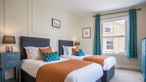 A hotel room with two beds, each with a headboard, white bedding, and orange throw blankets. There are bedside tables with lamps, a window with curtains, and framed pictures on the wall. - Sleeps 18 Central York 5★Townhouse Parking Cinema & Pool Table & Games Room (York)