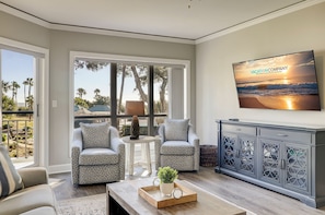 Un salon comprenant deux fauteuils à motifs, une table basse avec un plateau et une plante, un meuble avec des motifs décoratifs et un téléviseur affichant le texte « VACATIONCOMPANY ».