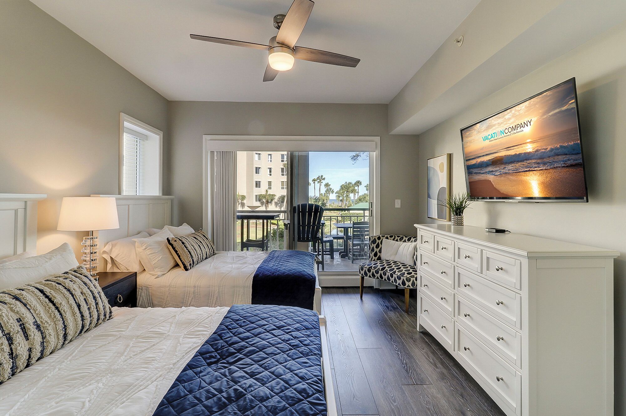 A hotel room with a bed, a desk, a chair, a TV, and a balcony with a view of palm trees and buildings.