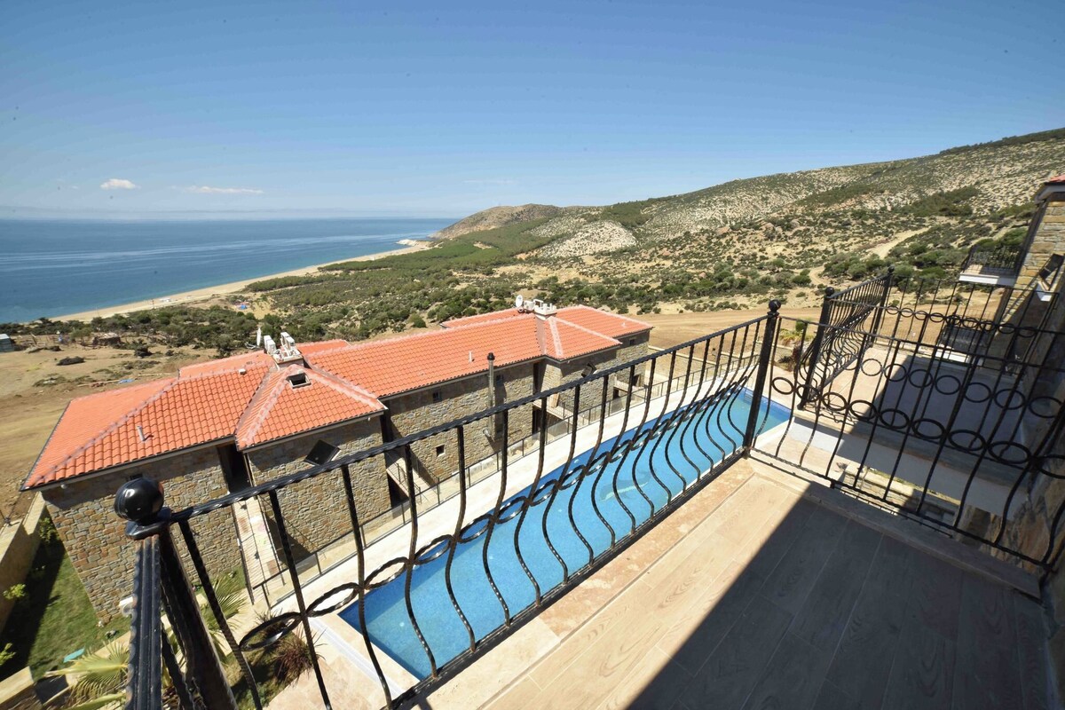 A balcony with a pool, overlooking a beach and hilly landscape.