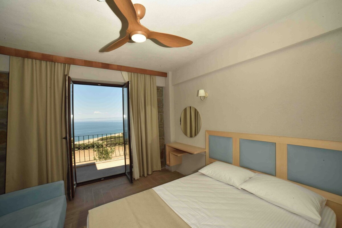 Habitación de hotel con cama, escritorio, ventilador de techo, vista al océano y balcón con cortinas.