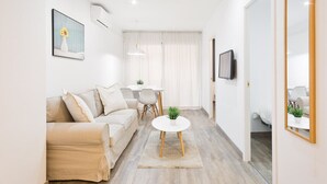 A modern living room with a beige sofa, a small round table with a plant, and a dining area with a white table and chairs. - GE1 (Barcelona)