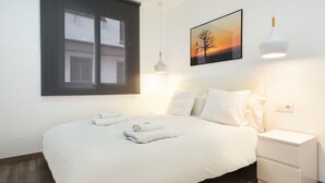 A neatly made bed with white linens and two towels, a bedside table with a lamp, and a window with blinds. - GE1 (Barcelona)