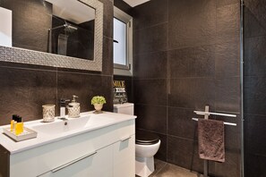 A modern bathroom with dark tiles, a white vanity, a mirror, a toilet, and a towel rack. - GE6 (Barcelona)