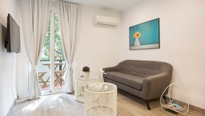 A modern living room with a grey sofa, a round glass coffee table, a white side table with a plant, and a white wire shelf. - G13 (Barcelona)