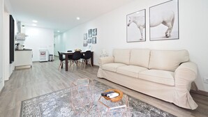A modern living room with a beige sofa, a coffee table, and a dining area with a table and chairs. There are framed pictures on the wall and a kitchen area in the background. - G14 (Barcelona)