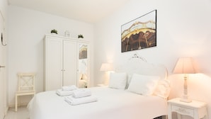 Une chambre à coucher blanche, avec un lit, deux tables de chevet dotées de lampes, une armoire blanche et un tableau encadré au mur.