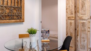 A glass table with a potted plant, a wooden bench, and a black chair in a room with a wooden door and a wall-mounted decorative piece. - G25 (Barcelona)