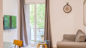 A living room with a glass table, a sofa, a clock, and a view of greenery through a window. - G25 (Barcelona)