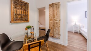 A room with a glass table, two black chairs, a wooden floor, a white wall with a decorative panel, and a door leading to another room. - G25 (Barcelona)